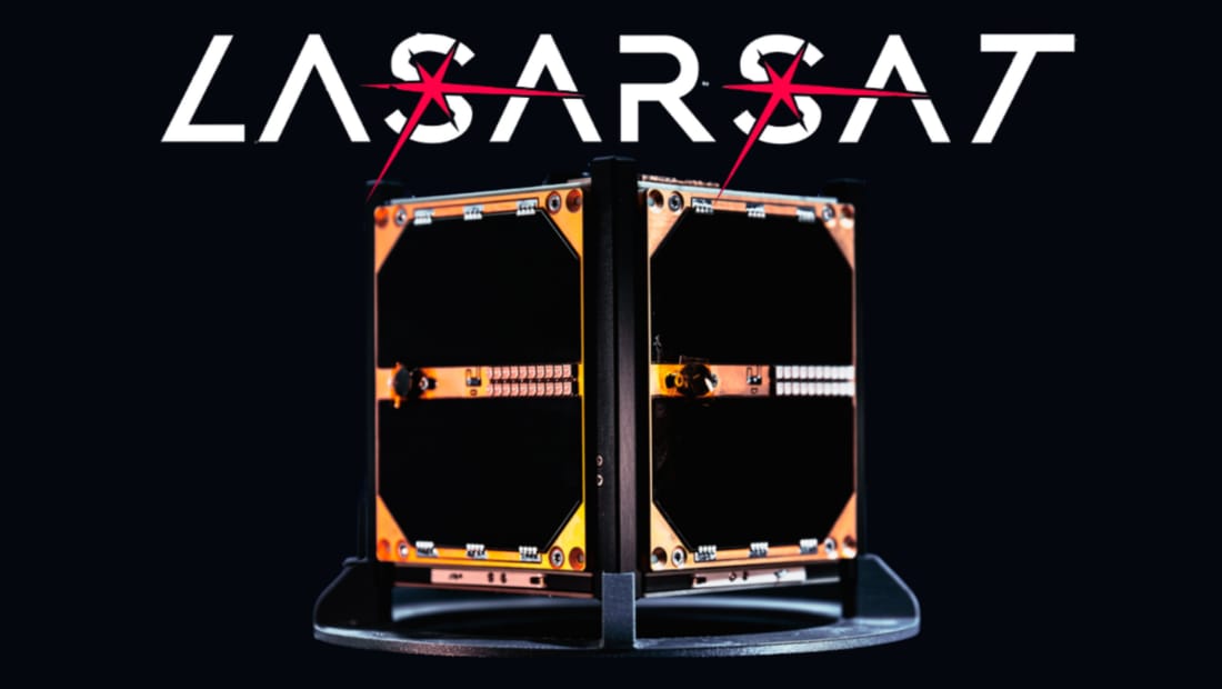 The LASARSAT CubeSat photographed on a stand against black, its gold-anodised structural frame holding dark solar panels with a row of small components across the middle. The LASARSAT wordmark above it is crossed by two red laser marks.